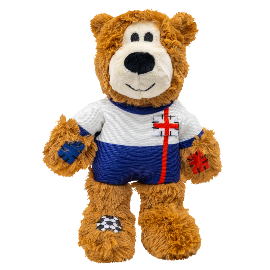 A front-facing studio product shot of the KONG Wild Knots England Bear dog toy against a white background. The brown plush bear is dressed in a white and blue football kit featuring a Red St George's Cross emblem on the chest. It includes decorative blue and red "stitched" patches on its arms and a football pattern on its right foot. The toy is designed with a durable plush exterior and a minimal stuffing interior to satisfy natural chewing instincts.