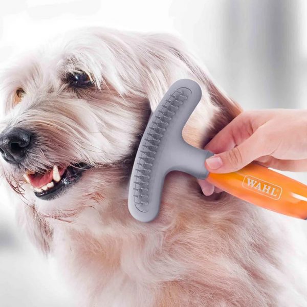 WAHL Double Row Rake Shedding Blade Lifestyle 1080px Close-up lifestyle photo of a person's hand using the WAHL grooming tool on a long-haired, white and tan terrier-cross dog. The grey rake head is buried gently into the dog's thick coat near its neck, demonstrating the brush in action. The translucent orange gel handle is held firmly, showing a comfortable grip during a home grooming session.