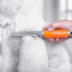 Close-up lifestyle photograph of a person's hand with manicured nails using the grey and orange WAHL dog grooming comb on a thick, curly, white-furred dog, such as a Poodle or Bichon Frise. The metal teeth of the comb are shown smoothing through the dense white coat to detangle the fur. The hand firmly grips the translucent orange gel handle, which clearly displays the silver WAHL logo