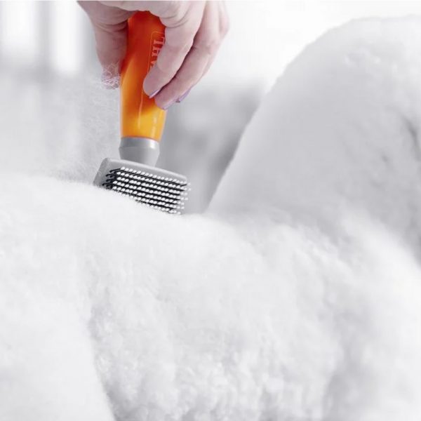 WAHL Nylon Slicker Brush Small Lifestyle2 1080px Close-up action shot of a hand using the orange-handled WAHL slicker brush to groom the thick, white curly coat of a dog. The fine white nylon bristles are shown moving through the fur to pull away loose hair