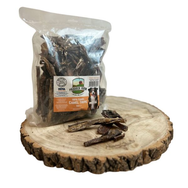 nova-dog-chews-paddock-farm-camel-tripe-1kg A 1kg clear plastic bag of Nova Dog Chews Paddock Farm 100% Natural Camel Tripe dog treats, standing upright on a rustic round wooden log slice against a clean white background. A few individual pieces of the dark brown, textured, air-dried camel tripe pieces are displayed neatly on the wood in front of the bag. The orange and cream product label features a Border Collie illustration and detailed nutritional information